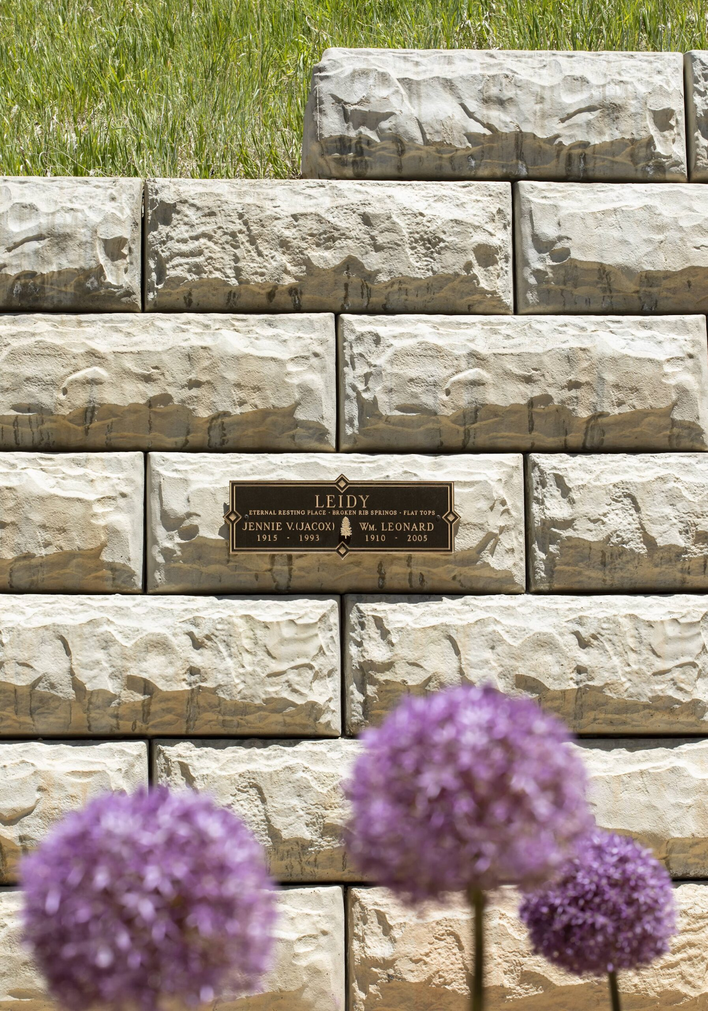 A stone wall with a plaque that reads "LEIDY" and "JENNIE V. 1915-1993, WM. LEONARD 1910-2005," with purple flowers in the foreground.