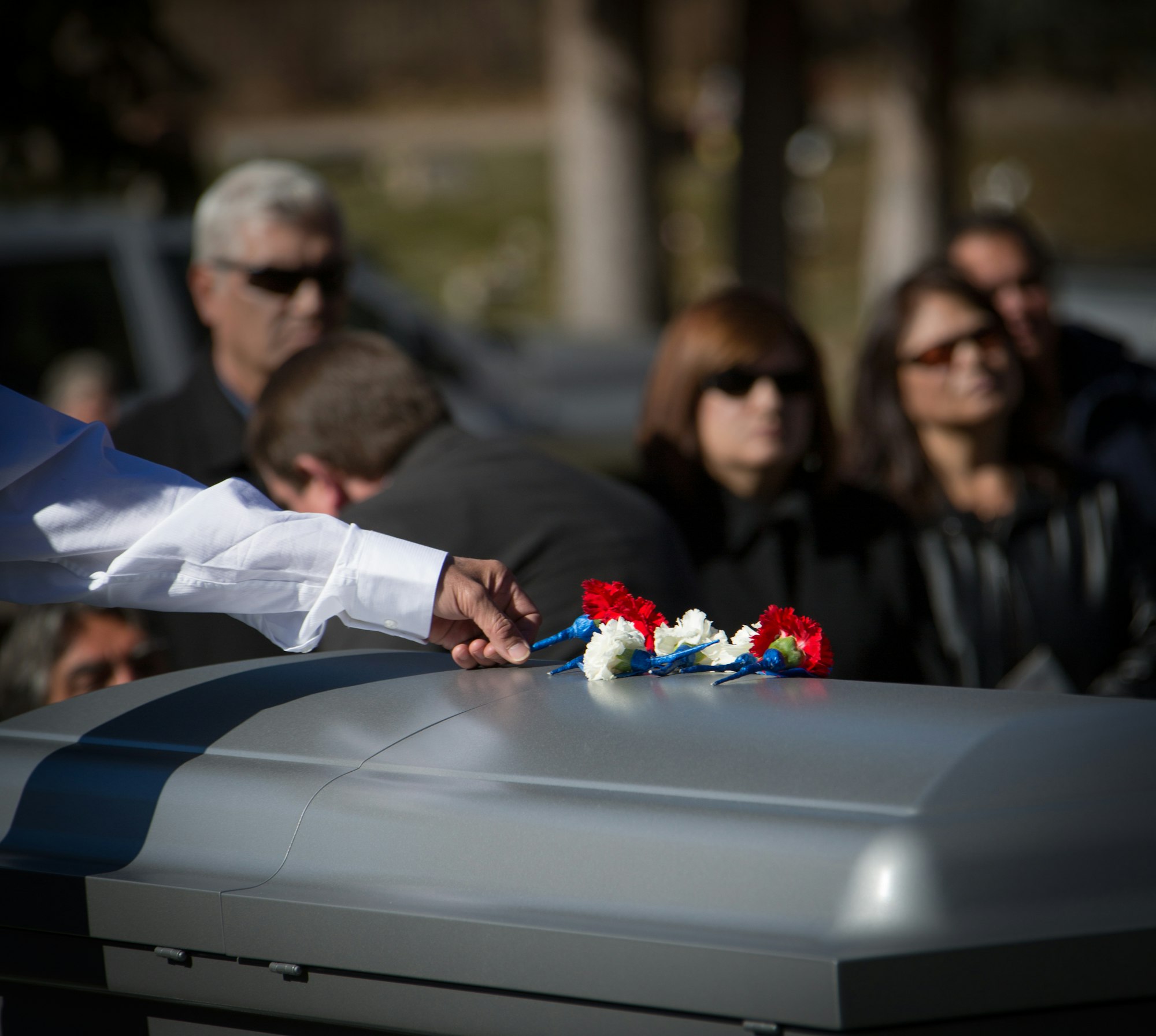 A hand places flowers on a casket at a funeral, with mourners in the background.