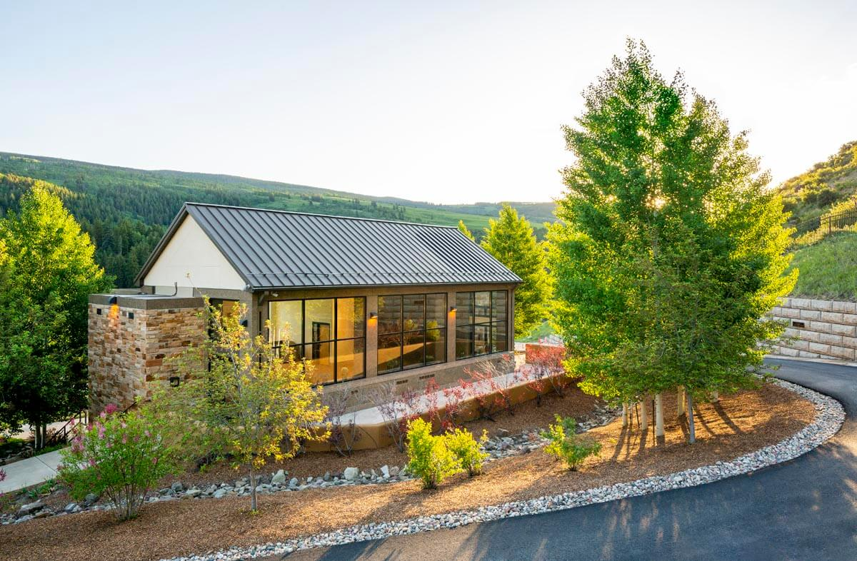 Modern house with large windows surrounded by trees and landscaped garden, set against a mountainous backdrop.
