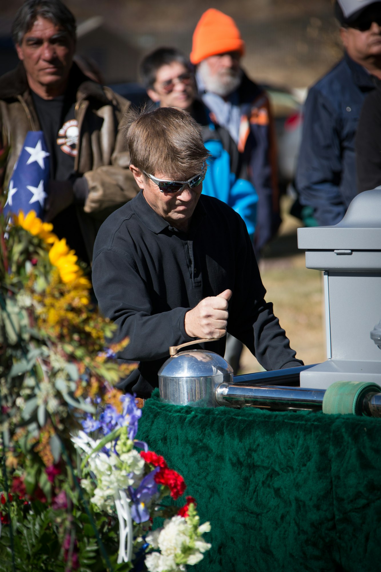 A man at a memorial service with flowers and people in the background, holding a silver container, possibly an urn.
