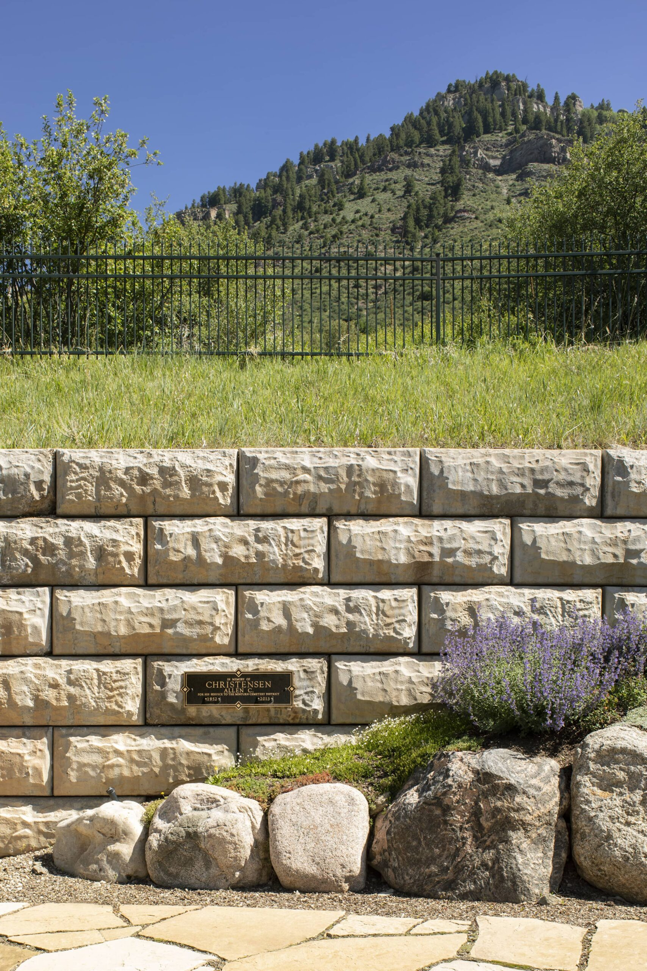 Stone wall with a plaque, a grassy area with a black fence, trees, and a mountain in the background under a clear blue sky.