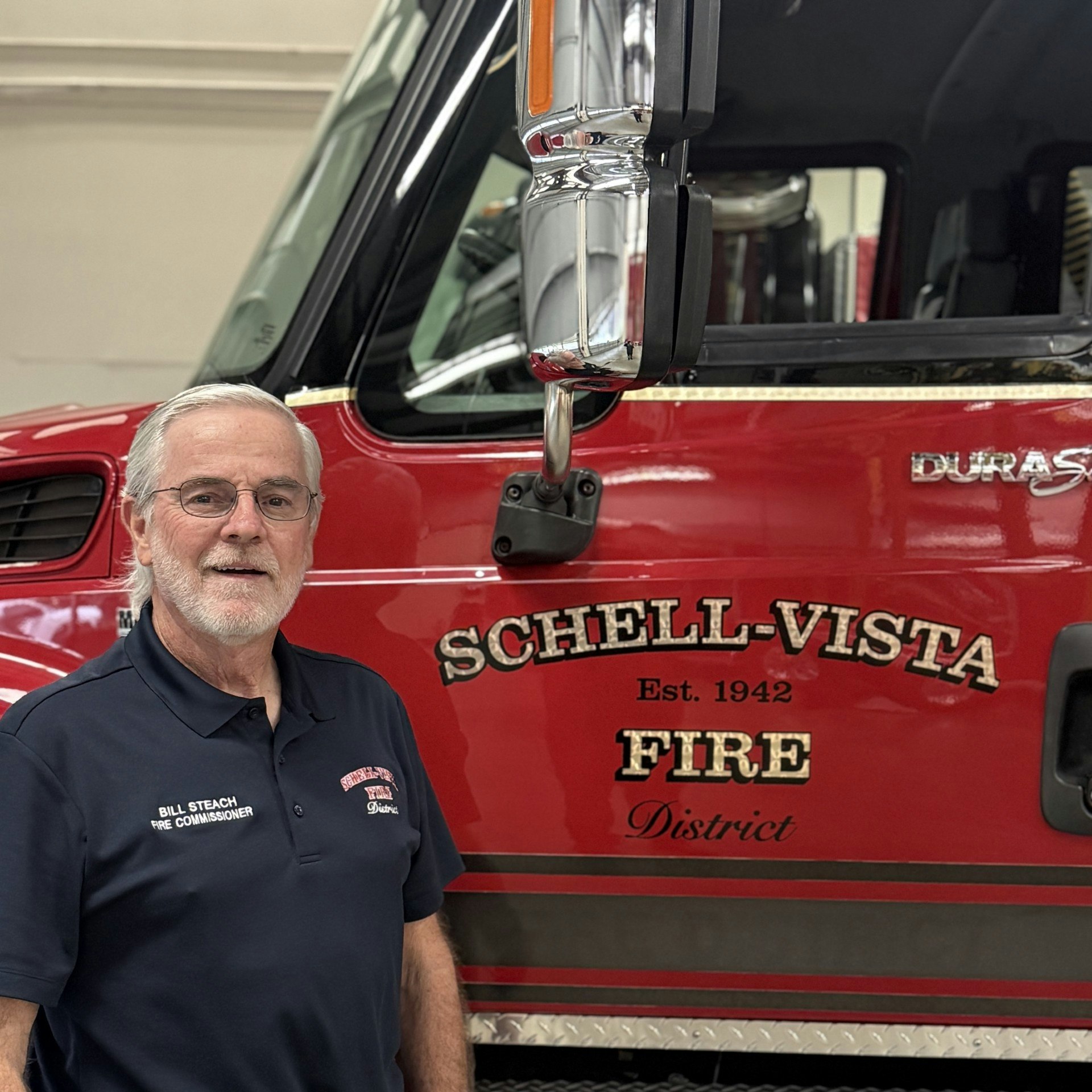 A man in a fire department shirt stands next to a red fire truck, displaying "Schell-Vista Fire District, Est. 1942."