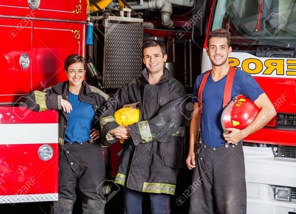 Three firefighters pose in front of a fire truck, wearing gear and smiling. One holds a helmet, another a fireman’s hat.