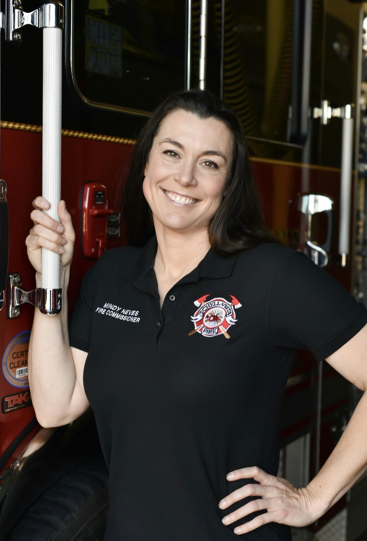 A smiling woman in a black polo shirt stands next to a fire truck, identified as a fire commissioner.