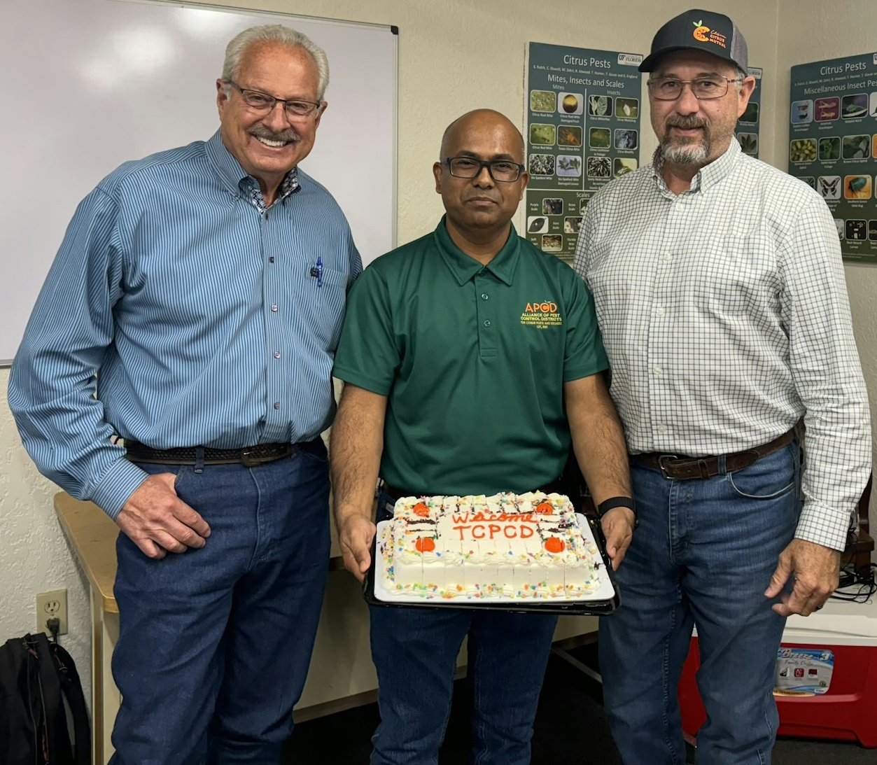 Three men smiling, holding a decorated cake with text, in a room with educational posters.