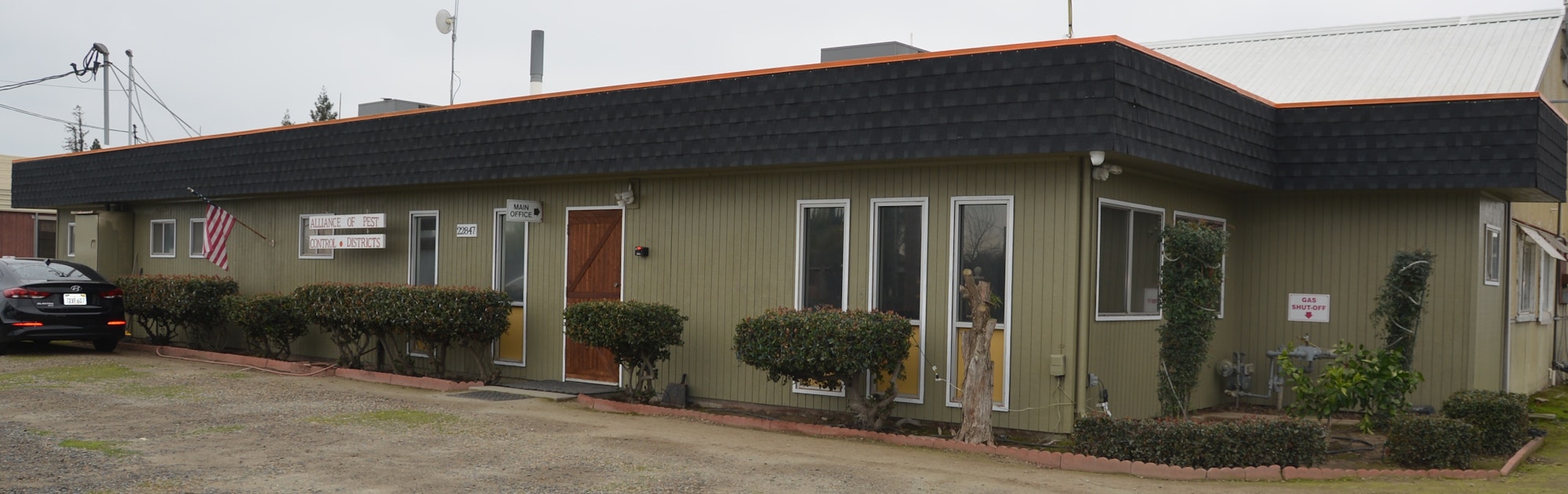 A single-story office building with a sign for "Alliance of Pest Control Districts," an American flag, and a parked car.
