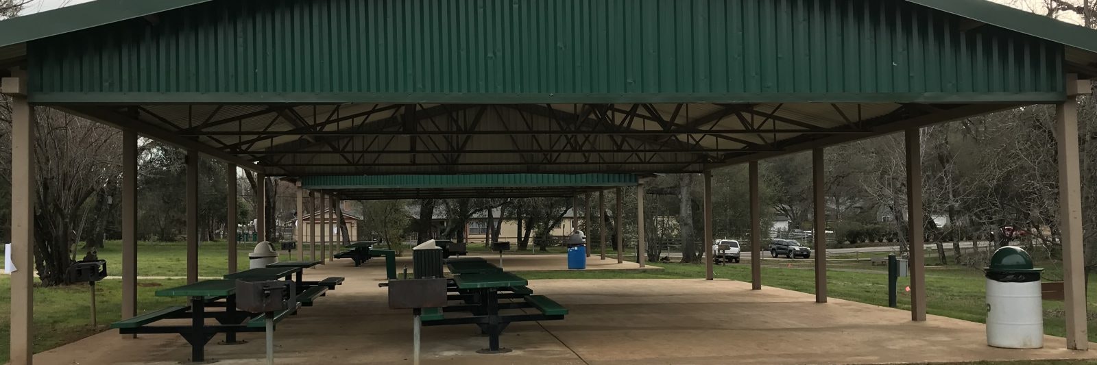 Covered picnic area with green tables, benches, grills, and trash cans in a park setting.