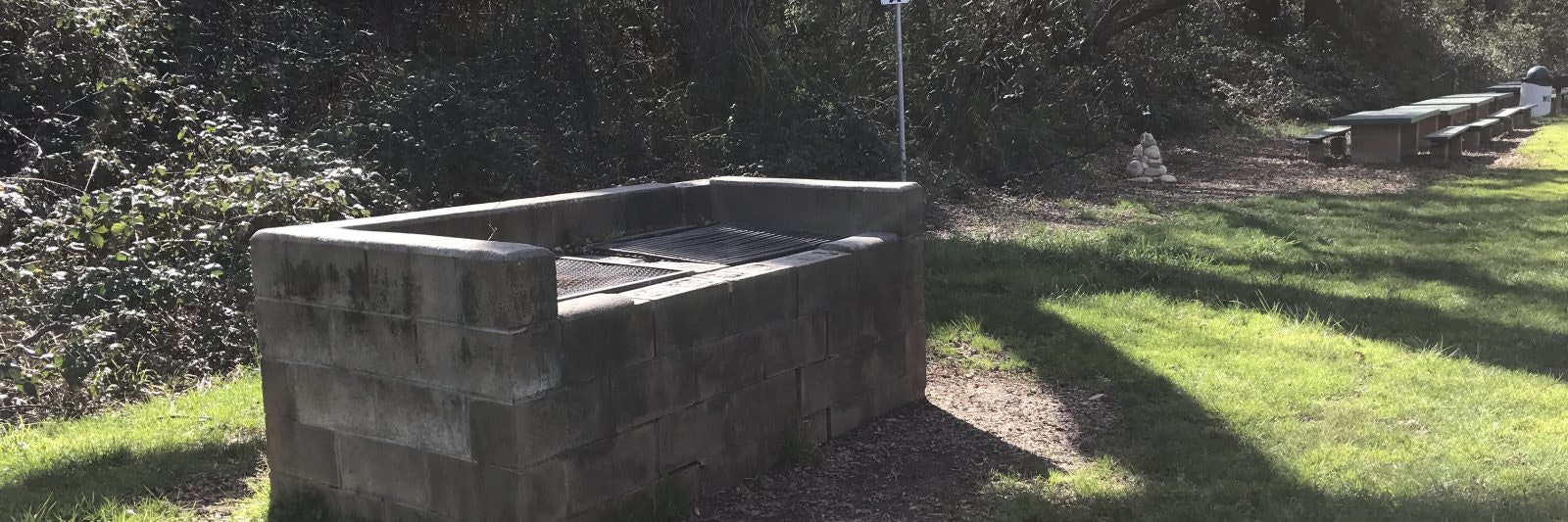A stone grill in a grassy area with picnic tables and a small rock pile nearby.