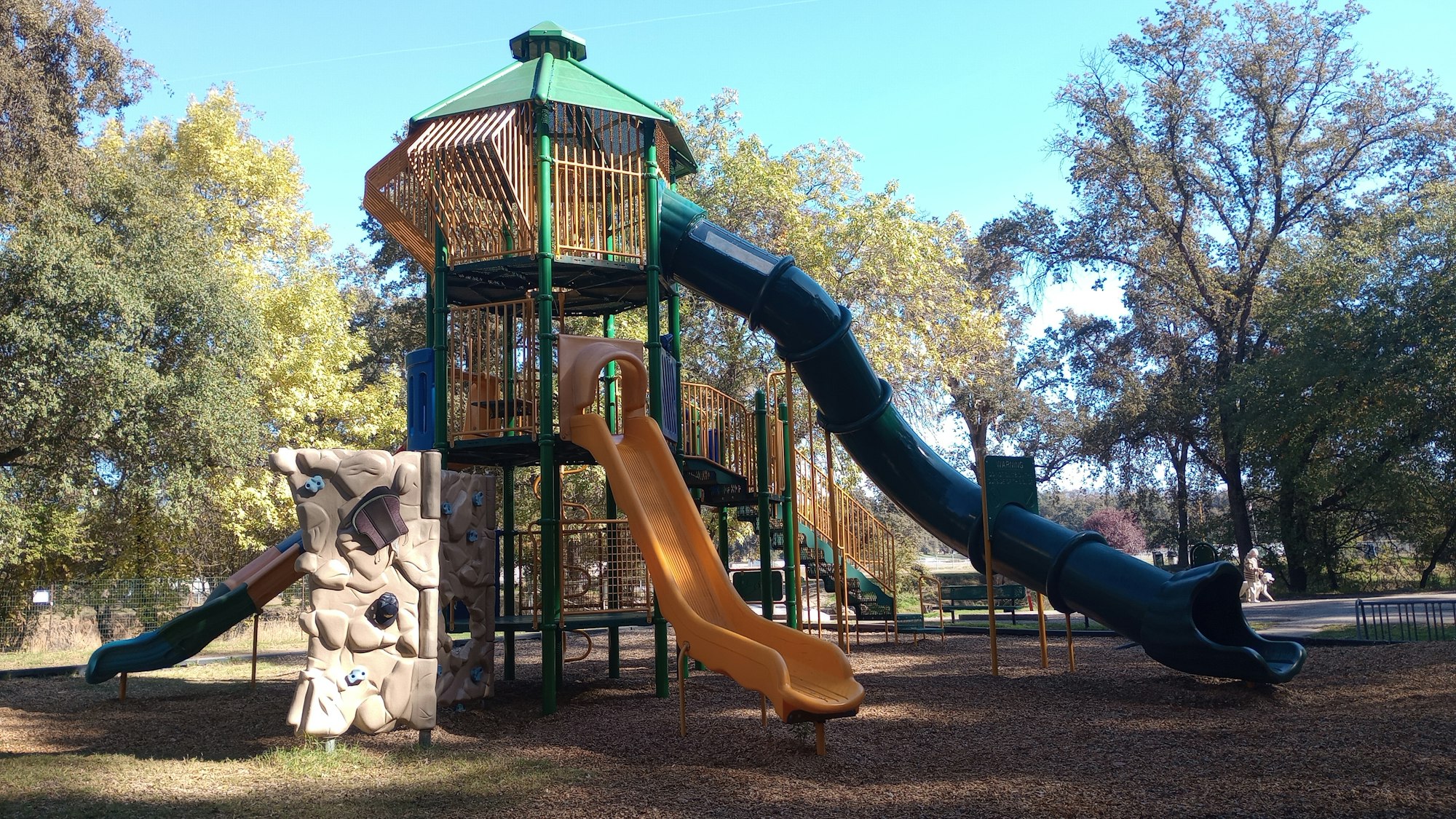 A playground with a large play structure featuring slides and climbing walls, set in a park with trees.