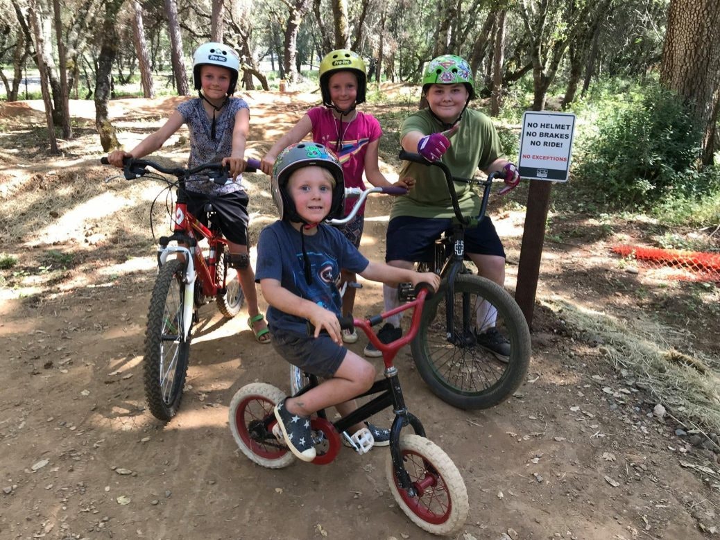 Four kids on bikes with helmets in a wooded area, near a sign saying "No helmet, no brakes, no ride! No exceptions."