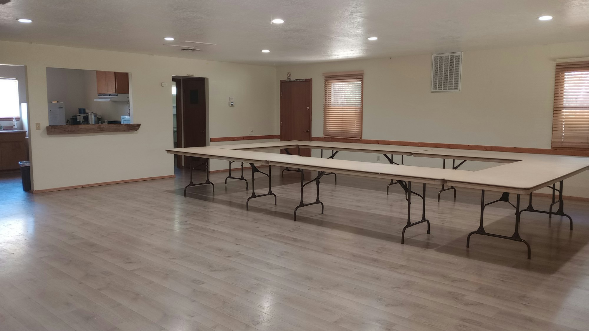 An empty room with U-shaped tables, wooden floor, and a kitchen area in the background.