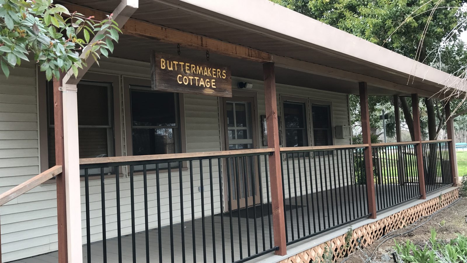 A cottage with a porch, sign reads "BUTTERMAKERS COTTAGE," surrounded by trees.