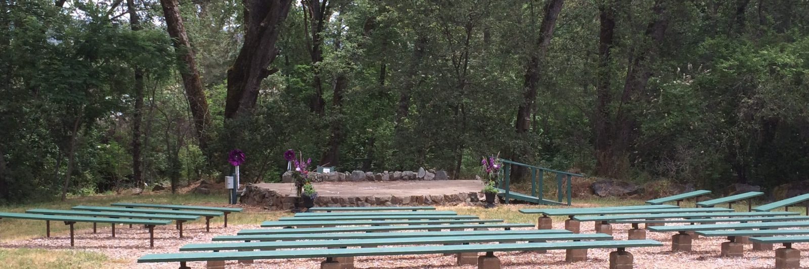 Outdoor amphitheater with green benches, surrounded by trees and featuring a small stage with purple floral decorations.