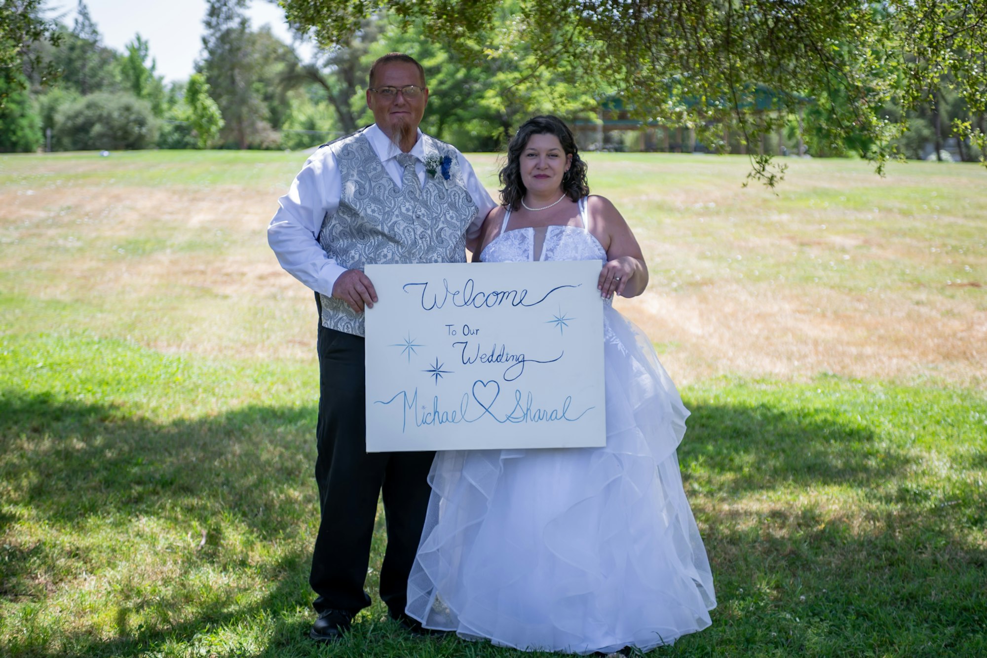A couple in wedding attire holds a "Welcome to Our Wedding" sign outside of the Buttermaker's Cottage.