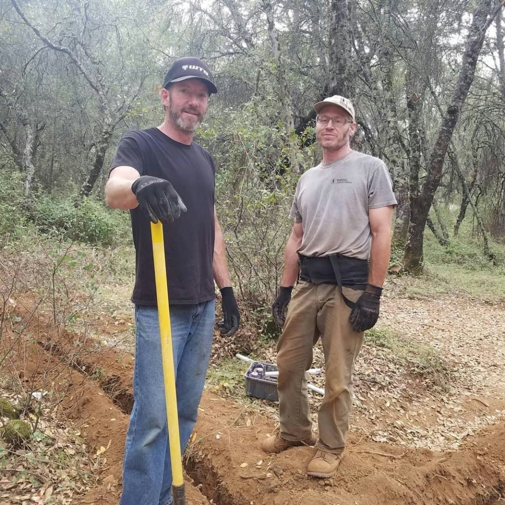 Two men in work attire standing in a forested area with a shovel and tools on the ground.
