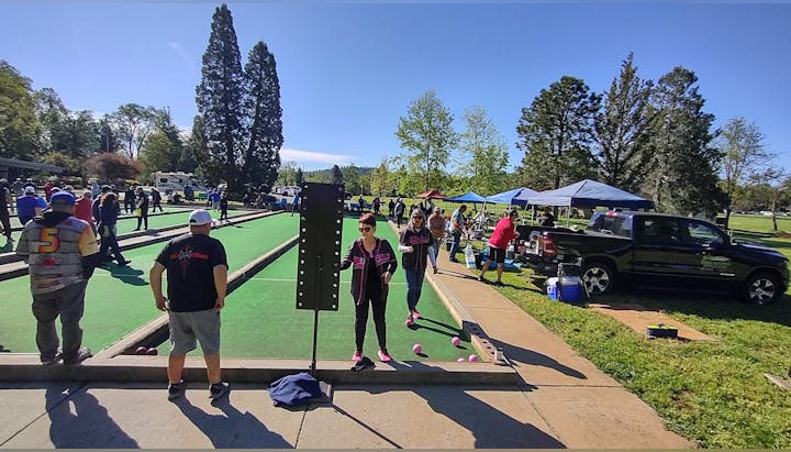 People are playing bocce on outdoor courts, with spectators and tents in a park setting.