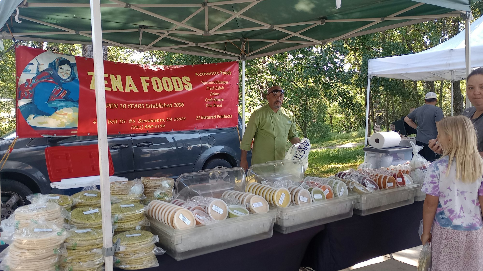 Outdoor food stall selling Mediterranean products like hummus and pita, with a vendor, banner, and customers.
