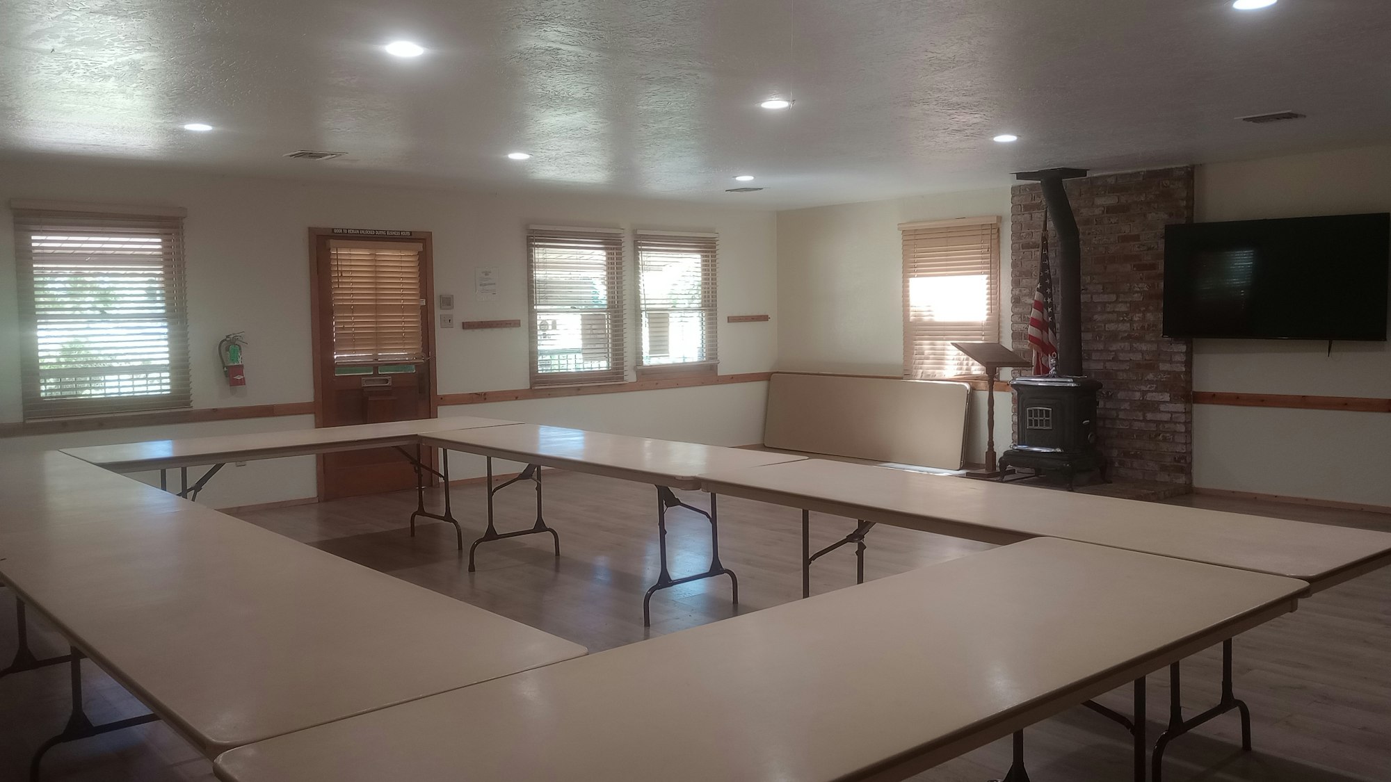 A meeting room with U-shaped tables, chairs, a brick wall, an American flag, a TV, and a wood stove.