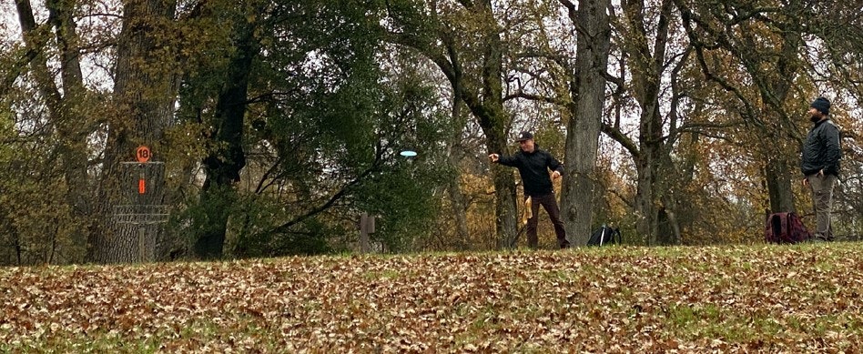 Two people playing disc golf in a wooded area, with one throwing a disc towards a basket.