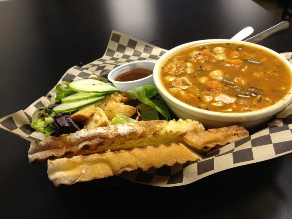 Bowl of vegetable soup, side salad with cucumbers and croutons, and crusty bread on checkered paper.
