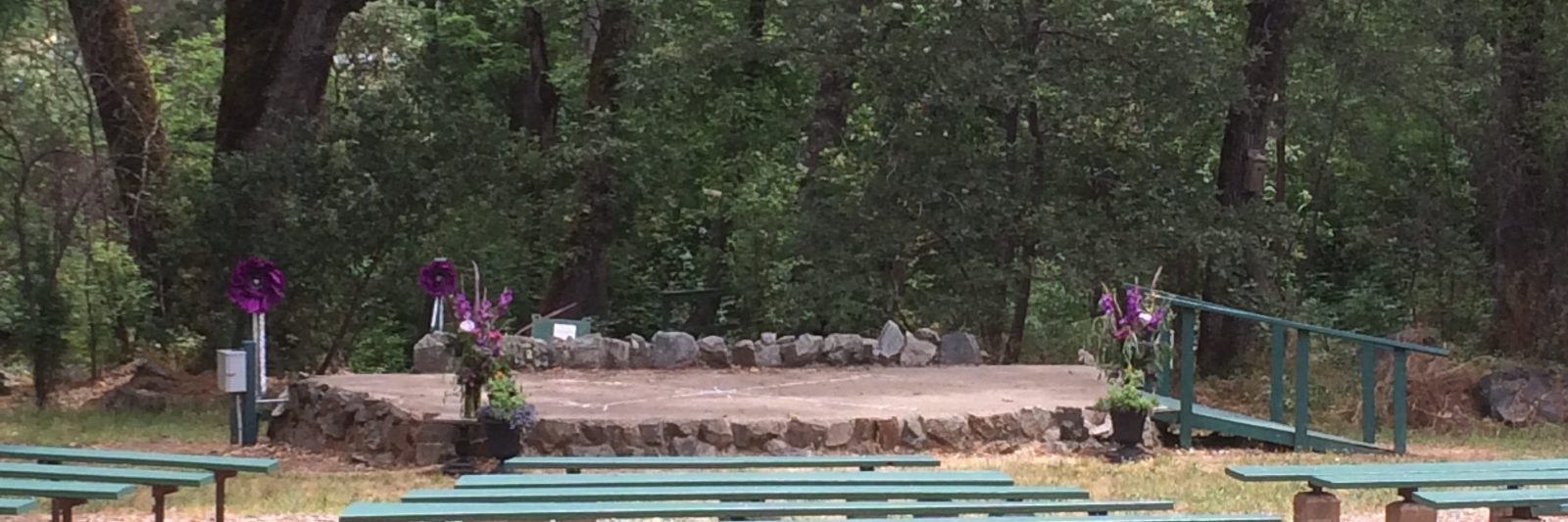 Outdoor stage with stone platform, surrounded by trees, and decorated with purple flowers. Green benches in the foreground.