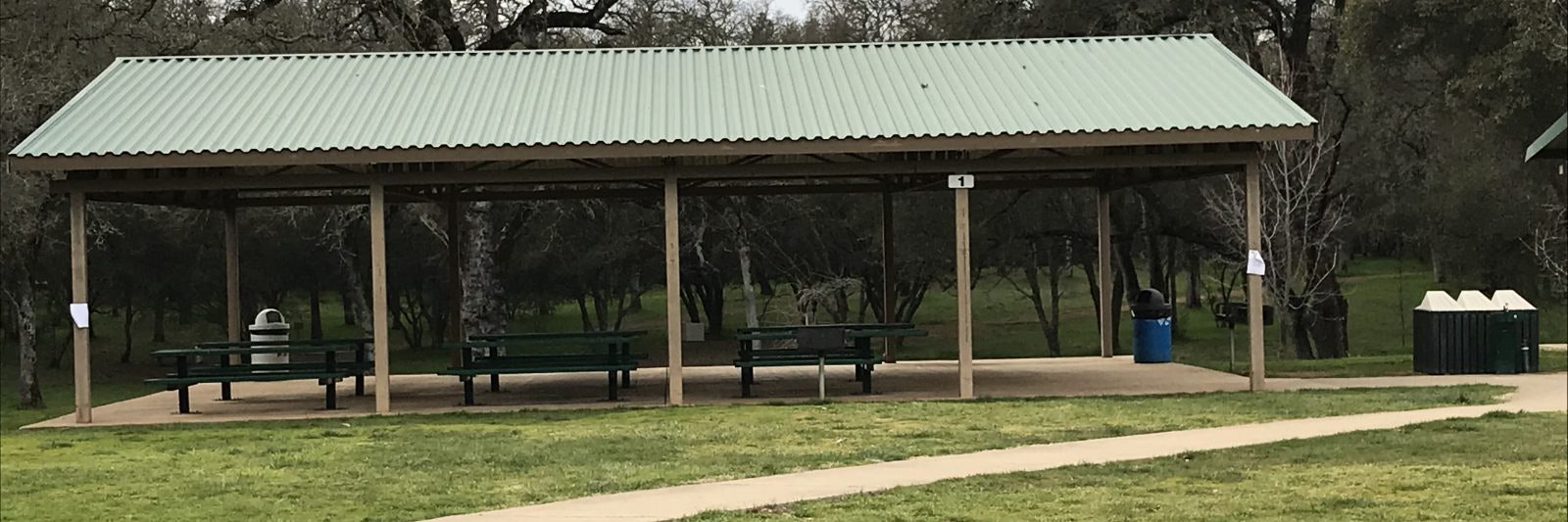 A park shelter with green roof, picnic tables underneath, surrounded by trees and grass.