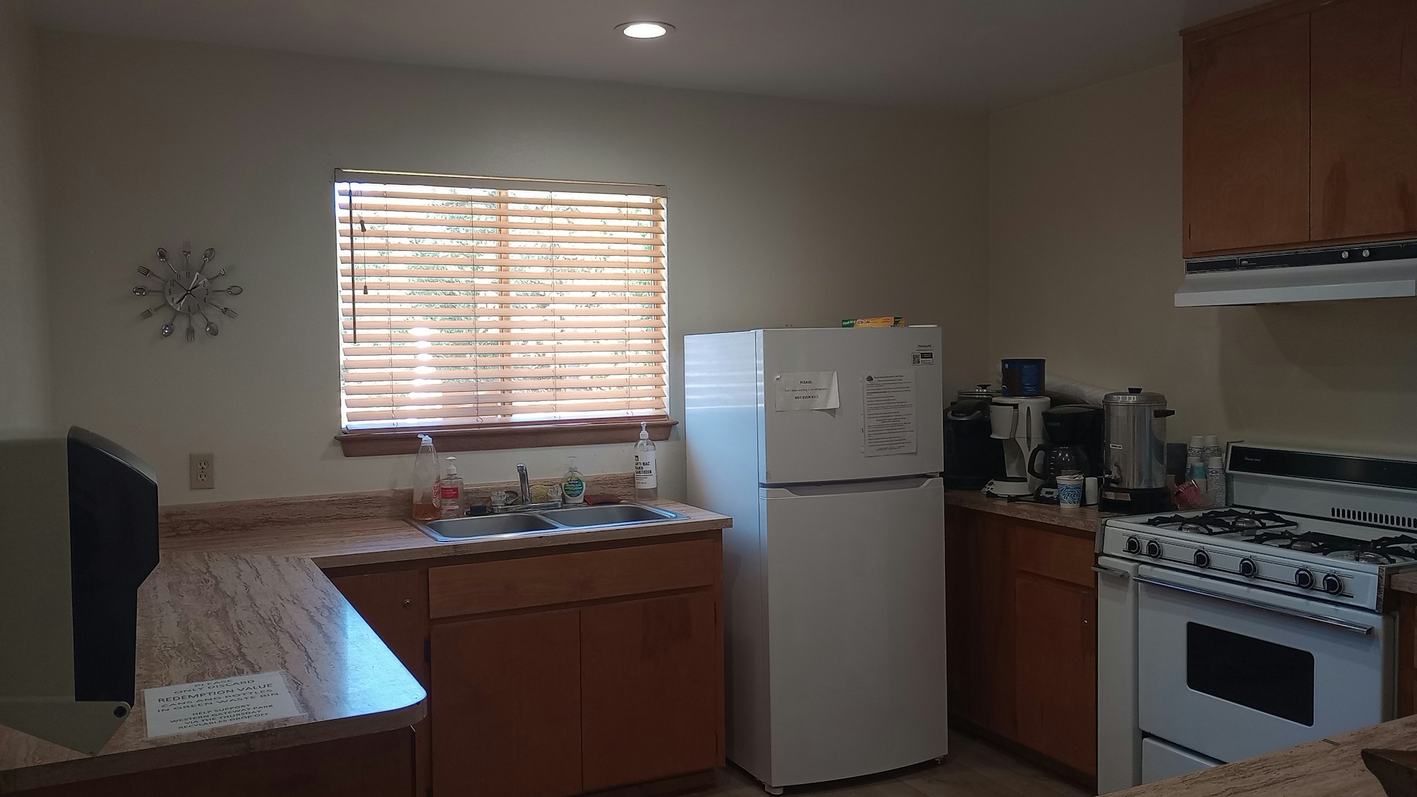 A kitchen with a refrigerator, stove, sink, wooden cabinets, and countertop appliances. A window with blinds and a wall clock are visible.