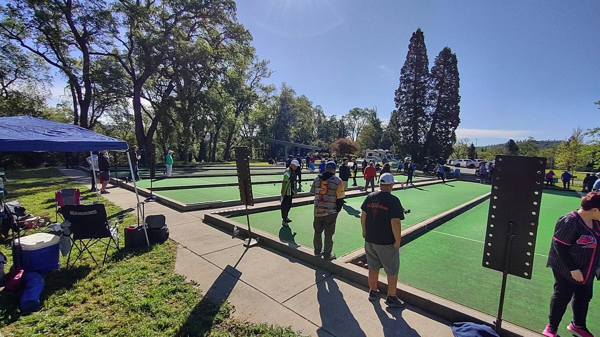 People playing bocce on outdoor courts with trees and a tent nearby.