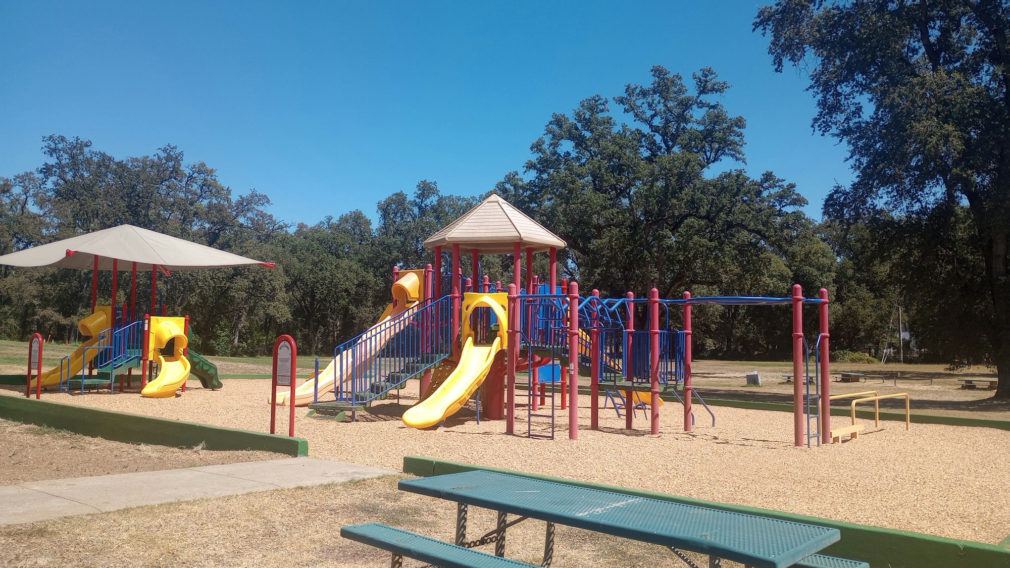 Colorful playground equipment with slides and climbing structures, surrounded by trees under a clear blue sky.