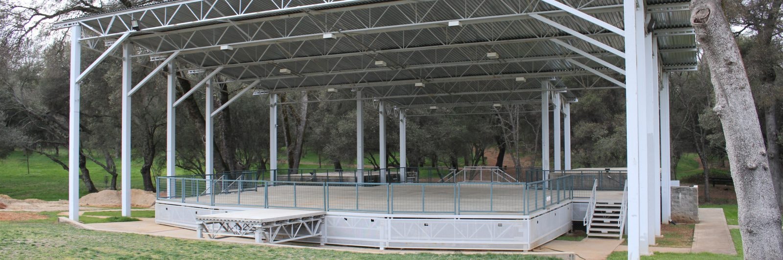 Open outdoor stage with metal roof and railings, situated in a park-like setting with trees in the background.