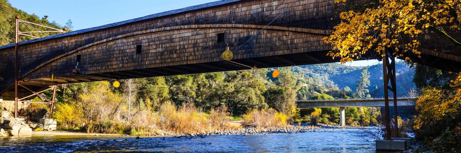 Wooden covered bridge over a river with autumn trees and a distant hill.