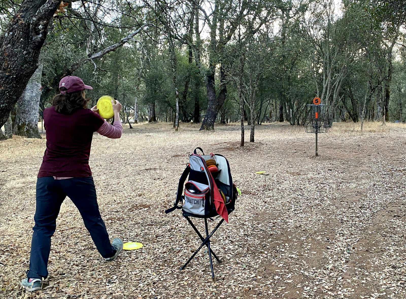 Person playing disc golf in a wooded area, aiming a yellow disc at a metal basket.
