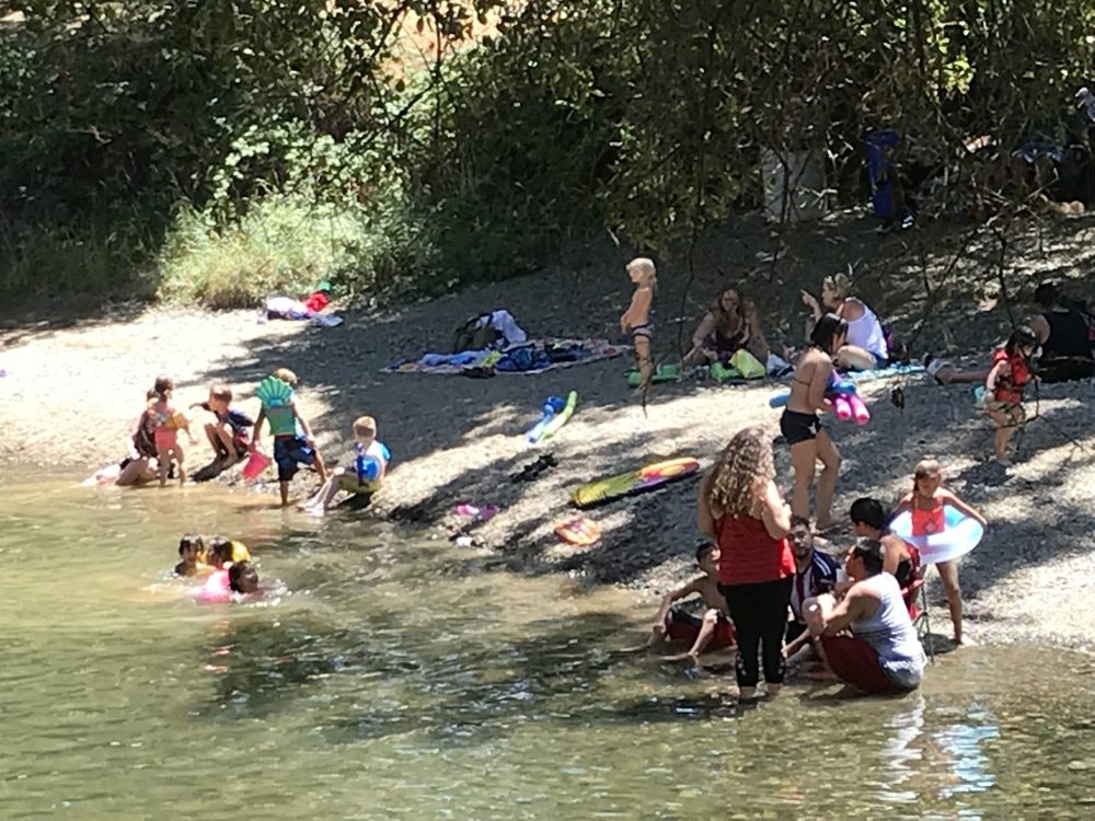 People enjoying a sunny day at a rocky lakeside, with children playing in the water and families relaxing on the shore.