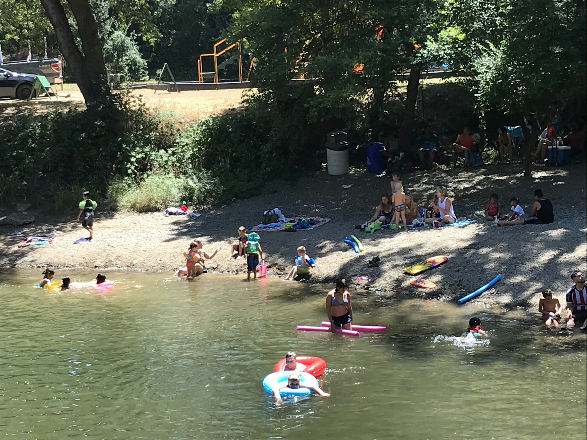 People enjoying a day at a lakeside beach, swimming and relaxing with inflatables and towels on the shore.
