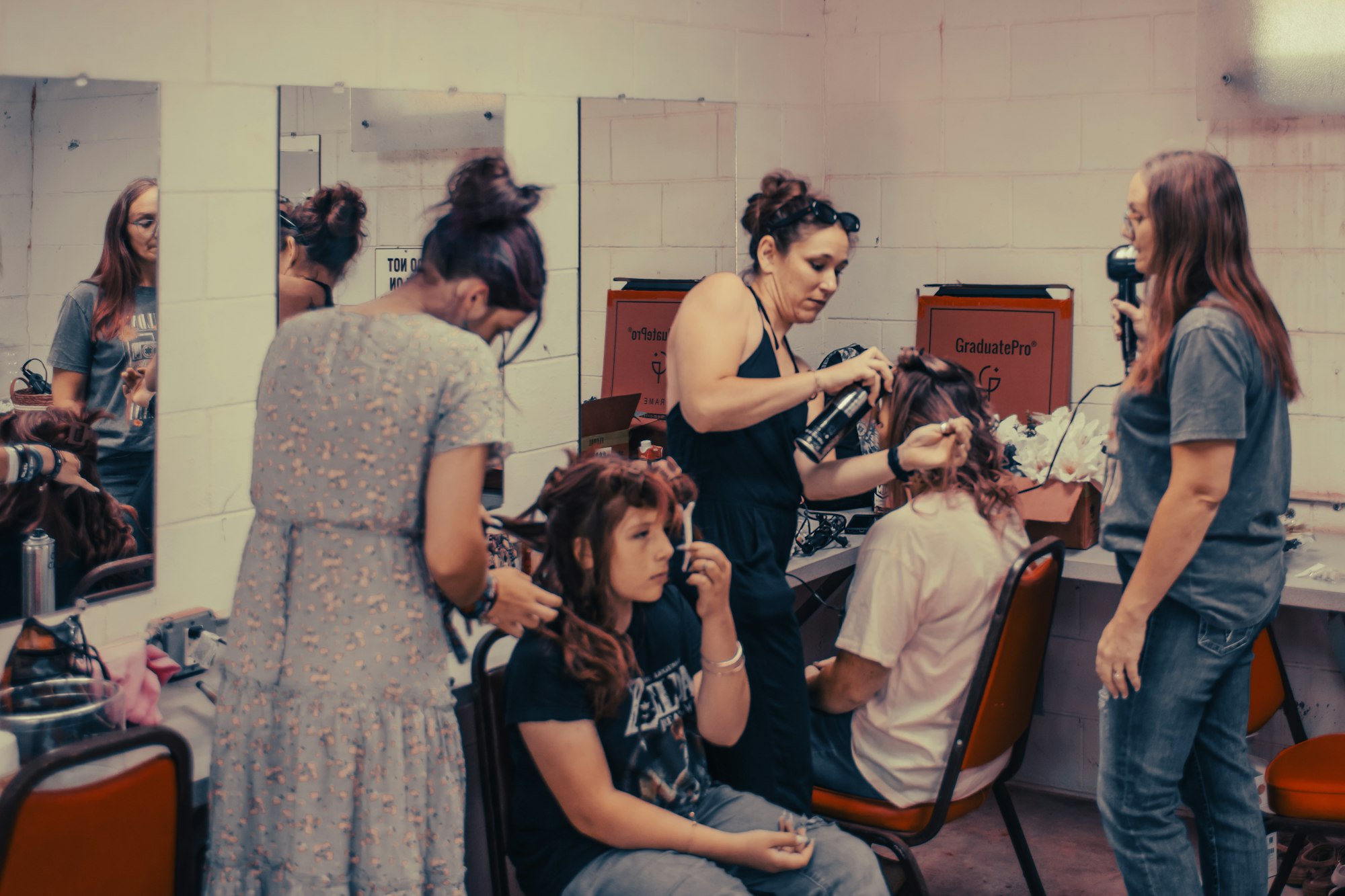 Two women are doing hair styling, using curlers and a hairdryer, on seated individuals in a Peirce Stage Pavilion green room.