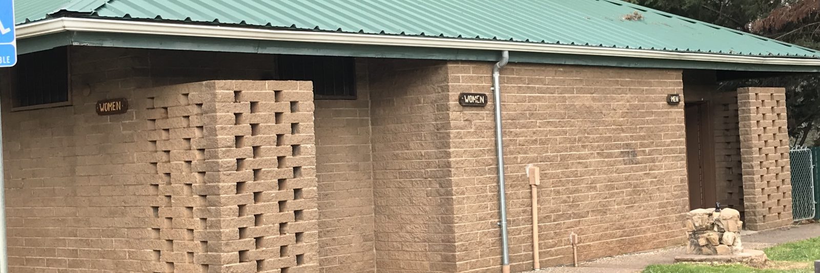 Brick building with signs for "Women" and "Men," indicating restrooms, under a green metal roof.