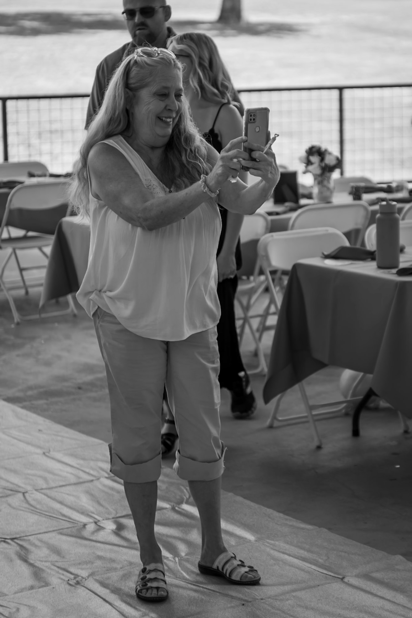 A woman taking a photo with her phone, surrounded by wedding guests and tables with flowers on the Peirce Stage Pavilion.