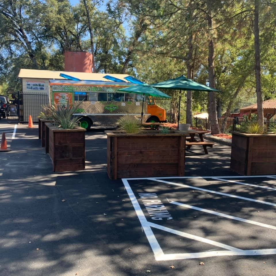 A food truck in a wooded area with picnic tables, large planters, and "No Parking" zone marked on the pavement.