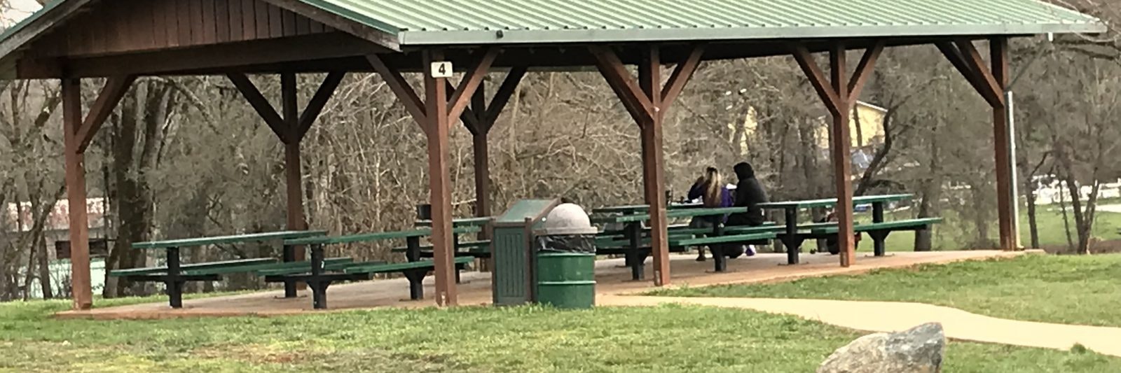 A park shelter with a green roof, picnic tables, a trash can, and two people sitting. Trees and grassy area in the background.