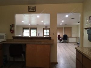 Room with wooden cabinets, a desk, chairs, and a microwave. It has windows, doors, and bright ceiling lights.