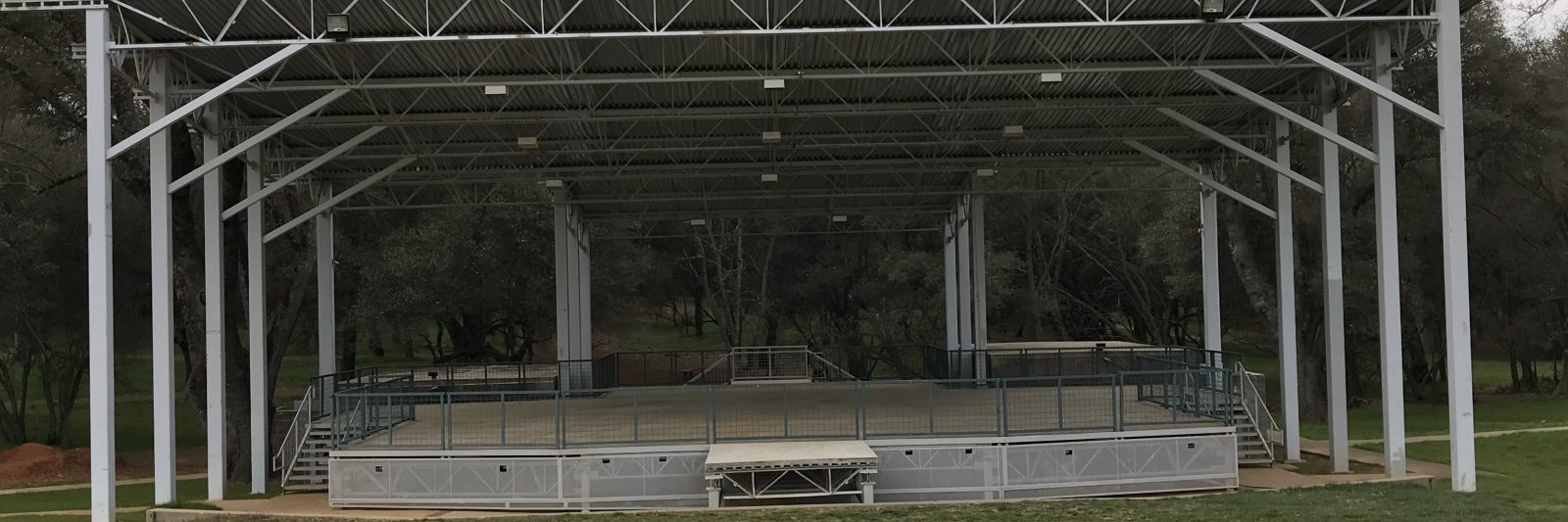 Outdoor stage with metal roof and surrounding railing, set in a grassy area with trees in the background.
