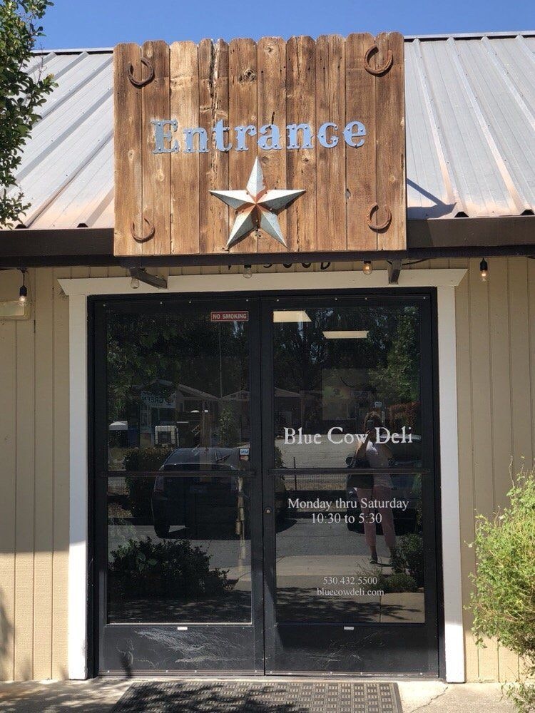Storefront with "Blue Cow Deli," entrance sign, star decoration, and operating hours displayed on glass doors.
