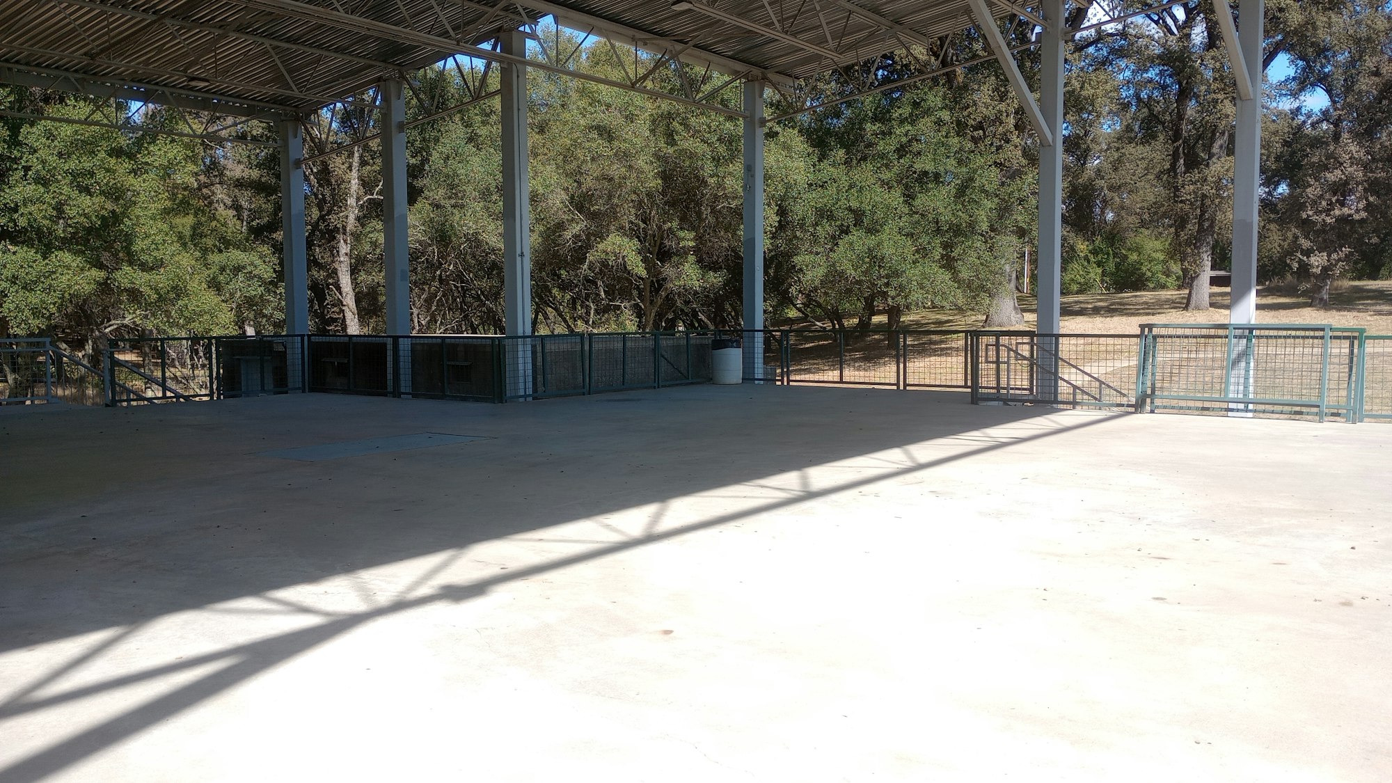 Open-air structure with metal beams, canopy roof, and fenced area, surrounded by trees.