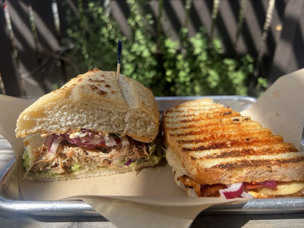 Two sandwiches on a tray: one with pulled meat and veggies, the other grilled with red onions and cheese. Served outdoors.