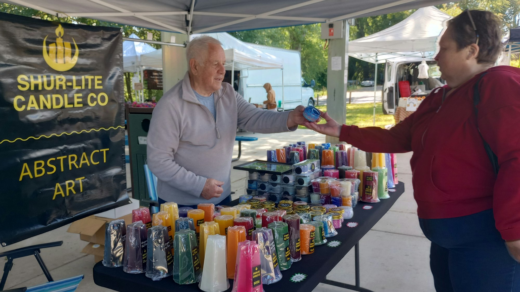 Two people at a candle stall, exchanging a candle at a market. Display of colorful candles.