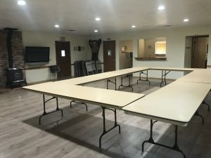 A U-shaped arrangement of tables in a room with a TV, folding chairs, and a brick stove.