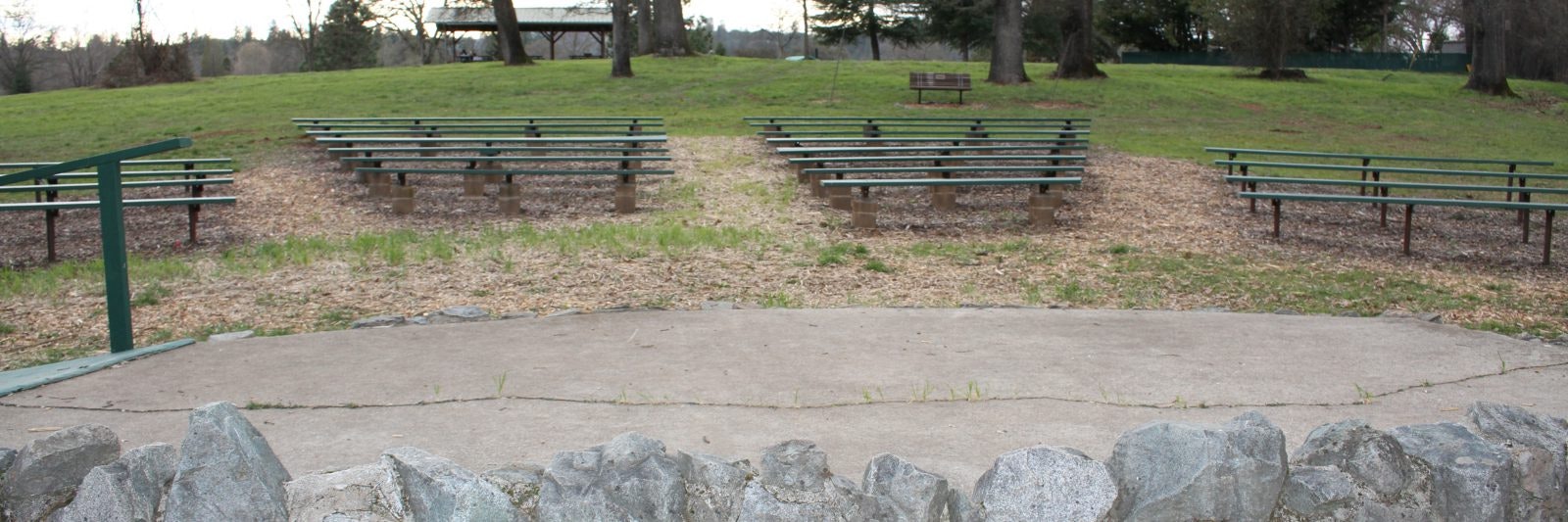 Outdoor seating area with benches facing a stone-framed stage, surrounded by grass and trees.