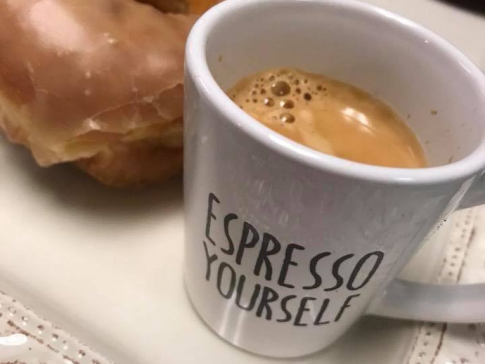 A mug with "Espresso Yourself" printed on it, filled with espresso, next to a glazed donut.