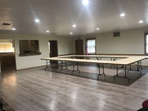 A large room with tables arranged in a U-shape, wooden floor, and bright lighting.
