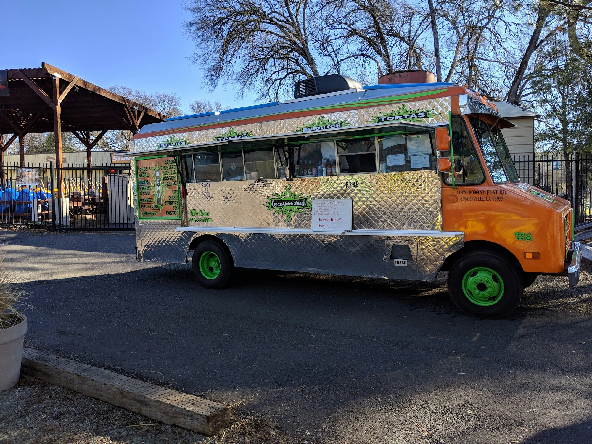 A shiny food truck with green and orange accents. It offers tacos, burritos, and tortas, parked near a wooden shelter.